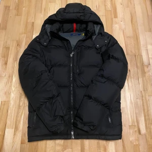 Svart dunjacka från Polo Ralph Lauren - Säljer en snygg svart dunjacka från Polo Ralph Lauren i storlek M. Jackan har en dragkedja framtill och en mysig huva. Perfekt för kalla vinterdagar. Den är i mycket bra skick och kommer i originalförpackning. Ett utskrivet kvitto kommer även med!