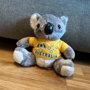 Gosedjur Koala med gul tröja - Söt koala plyschleksak klädd i en gul tröja med texten 'Australia' och stjärnor. Tillverkad av 100% polyesterfiber. Perfekt som souvenir eller present.