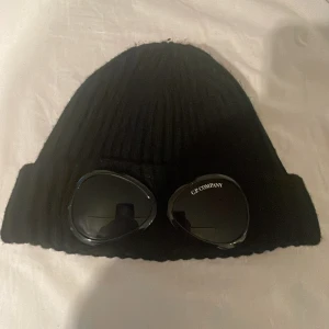 Svart mössa från C.P. Company - Snygg svart ribbad mössa från C.P. Company. Perfekt för höst och vinter med en cool detalj av glasögon framtill. Den är i mycket bra skick och passar perfekt för att hålla dig varm och stilren under kalla dagar.