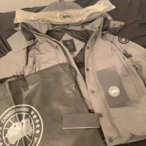 Grå dunjacka från Canada Goose - Säljer en stilren grå dunjacka från Canada Goose. Jackan har en klassisk design med knappar och dragkedja, samt en broderad logotyp på ärmen. Perfekt för kalla vinterdagar.