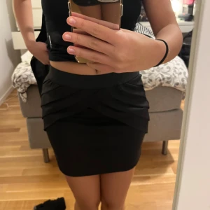 Svart kortkjol med volangdetaljer - Snygg svart kortkjol med volangdetaljer framtill. Kjolen har en tight passform och är perfekt för en stilren look. Den är enkel att matcha med olika toppar och accessoarer.