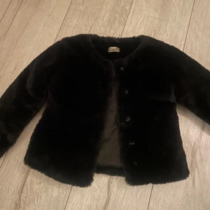 Svart pälsjacka från DJ Dutch Jeans - Säljer en stilren svart barn pälsjacka från DJ Dutch Jeans. Jackan har en klassisk design med knappar framtill och är perfekt för att hålla sig varm under kyliga dagar. Den är långärmad och har en mjuk och fluffig känsla. 3-5 år