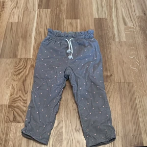 Grå prickiga byxor från H&M - Säljer ett par grå byxor från H&M med söta rosa prickar. Byxorna har en elastisk midja med snörning för extra komfort. Perfekta för en lekfull och bekväm stil.