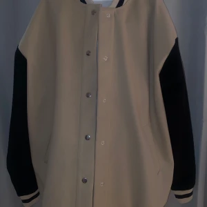 Beige och svart jacka med text - Snygg beige och svart jacka med texten 'AUTHENTIC' på ryggen. Jackan har svarta ärmar och knappar framtill. Perfekt för en cool och avslappnad stil.