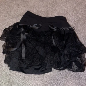 Svart kjol med spets och rosetter - Säljer en superfin svart kjol med volanger och spetsdetaljer. Den har två söta rosetter framtill som ger en extra touch. Det finns inga skador eller fläckar och är använd endast en gång!🫶🏻 säljer på grund av att den inte används🤍 