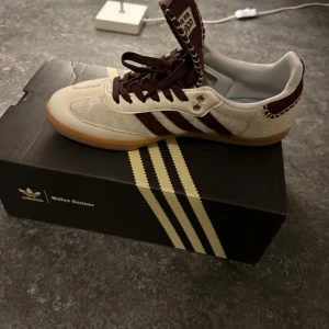 Wales bonner Sambas  i beige och vinröd - Säljer ett par snygga Wales Bonner  Sambas i vit/beige med vinröda detaljer som jag fick som present. Skorna har en klassisk design med snörning och en bekväm passform. Perfekta för både vardag och fest! De är i mycket bra skick och kommer i originalkartong. Pris kan diskuteras vid snabbaffär