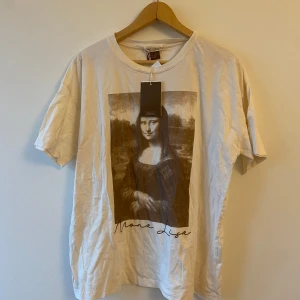 Vit t-shirt med Mona Lisa-tryck - Säljer en vit t-shirt med ett stort tryck av Mona Lisa på framsidan. T-shirten har en avslappnad passform och är kortärmad, perfekt för vardagsbruk. Materialet känns mjukt och bekvämt, och den har en rund halsringning. Perfekt för konstälskare eller som en cool statement-piece!