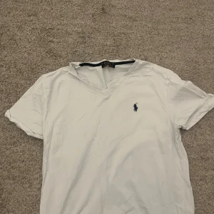 Vit t-shirt från Ralph Lauren - Säljer en klassisk vit t-shirt från Ralph Lauren med v-ringning och korta ärmar. Den har en broderad logga på bröstet. Perfekt för en stilren och avslappnad look. Den är bara ringlig man kan lösa det där enkelt med en strykjärn 