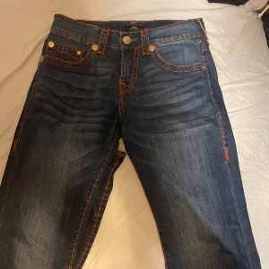 Blå jeans från True Religion - Säljer ett par snygga blå jeans från True Religion i modellen Ricky Relaxed Straight. De har coola orangea sömmar och klassiska femficksdesign. Perfekta för en avslappnad stil med en touch av edge. Passar både till vardags och fest!