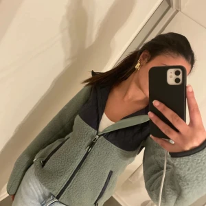 Grön fleecejacka från CRW - Säljer en supermysig grön fleecejacka från CRW i storlek XS. Jackan har en dragkedja framtill och är perfekt för kyliga dagar. Den är i nytt skick och har en snygg kontrast med mörkare detaljer vid dragkedjan och ärmsluten. Perfekt för höst och vår!