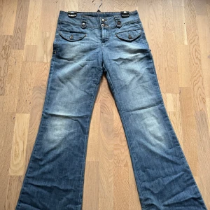 Blå lågmidjade bootcut jeans - Säljer ett par snygga blå bootcut jeans i bra skick, förutom lite slitage längst ner. De har en lågmidjade och coola knapplösningar fram och är perfekt för någon som har lite längre. Jag säljer jeansen då de tyvärr inte passar mig i storlek eller i längden eftersom att de är så pass långa på mig. Skriv gärna om ni har några frågor💗