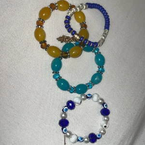 Färgglada armband med pärlor och berlocker alla armband kostar 15 kr - Säljer ett set med fyra armband i olika färger och stilar. Ett armband har blå och vita pärlor med en handberlock, ett annat har blå och vita pärlor med ögonmotiv. De andra två har stora pärlor i gult och turkost med glittriga detaljer. Perfekt för att lägga till en färgklick i din outfit!