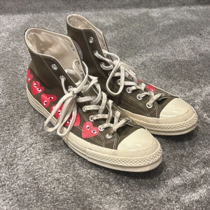 CDG CONVERSE - Snygga höga sneakers från Converse i olivgrön färg med röda hjärtan på sidan. Skorna har klassisk snörning och vit sula. 