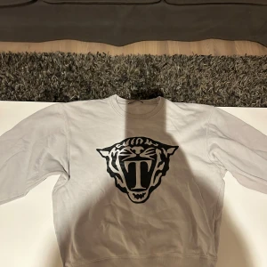 Grå sweatshirt med tryck Tiger of sweden - Säljer en grå sweatshirt med ett stort svart tryck av ett djurhuvud på framsidan. Tröjan har långa ärmar och en rund halsringning. Perfekt för en avslappnad stil.