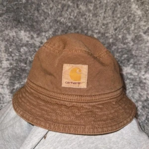Brun bucket hat från Carhartt - Snygg brun bucket hat från Carhartt i bomull. Perfekt för en avslappnad stil med klassisk logga framtill. Passar bra till både vår och sommar.