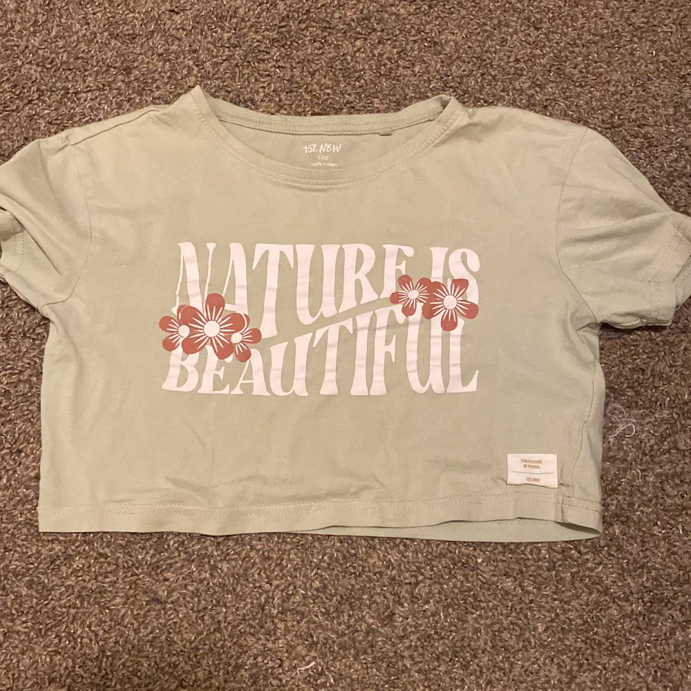 Säljer en superfin grön croppad t-shirt från 157 New. Den har ett tryck med texten 'Nature is Beautiful' och söta blommor i rosa och vitt. Perfekt för sommardagar! Gjord i 100% bomull, så den är riktigt skön att ha på sig. Passar perfekt till ett par jeans eller shorts.. Toppar.