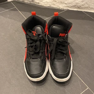 Svarta och röda sneakers från Puma - Snygga svarta och röda sneakers från Puma med hög ankel och snörning. Skorna har en vit sula och Puma-loggan på sidan och baksidan. Perfekta för en sportig look.