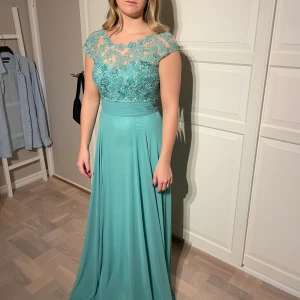 Turkos långklänning med spetsdetaljer - Elegant turkos långklänning med vackra spetsdetaljer på överdelen och korta ärmar. Klänningen har en figursydd midja och ett flödande kjolparti som ger en fin silhuett. Perfekt för speciella tillfällen.