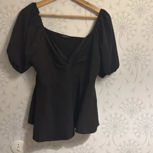 Svart blus med puffärmar - Snygg svart blus med puffiga kortärmar och v-ringning. Den har en smockad rygg och en lätt utsvängd nederdel som ger en fin siluett. Perfekt för en stilren look.