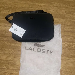 Svart axelväska från Lacoste - Snygg svart axelväska från Lacoste med justerbar rem och dragkedja. Perfekt för att bära dina dagliga nödvändigheter med stil. Väskan har en minimalistisk design med Lacoste-loggan diskret placerad på framsidan.