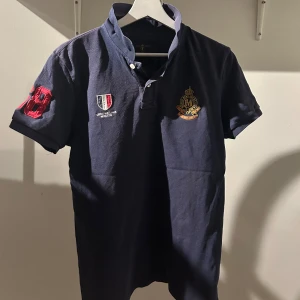 Mörkblå pikétröja från Morris - Snygg mörkblå pikétröja från Morris med broderade emblem på bröstet och ärmen. Tröjan har en klassisk krage och korta ärmar, perfekt för en stilren look. Märkesnamnet är broderat på kragen baktill. 