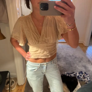 Beige omlottopp - Säljer en beige omlottopp med korta ärmar och v-ringad design. Toppen är croppad och har en luftig passform