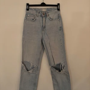 Ljusa jeans med hål från Gina Tricot - Snygga ljusa jeans från Gina Tricot med slitningar på knäna. De har en klassisk femficksdesign och knappgylf. Storlek 34.