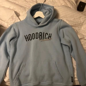 Ljusblå hoodie från Hoodrich - Säljer en ljusblå hoodie från Hoodrich med svart text på bröstet. Den har en klassisk känguruficka och en bekväm huva. Perfekt för en avslappnad stil. Sitter som xs ungefär 