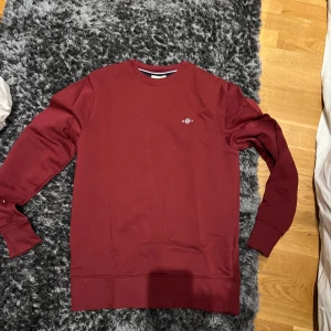 Röd sweatshirt från Gant - Snygg röd sweatshirt från Gant med rund hals och långa ärmar. Perfekt för en avslappnad stil. Den har en liten logga på bröstet som ger en stilren touch. Såklart billigare vid snabb affär!