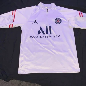 Psg tracksuit tröja - Snygg vit långärmad fotbollströja från Jordan i samarbete med PSG. Tröjan har röda och svarta detaljer samt texten 'ACCOR LIVE LIMITLESS' och 'VISIT RWANDA'. Perfekt för fotbollsfans som vill ha stil både på och utanför planen.