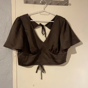 Brun omlottopp med knytband - Snygg brun omlottopp med korta ärmar och knytband i ryggen. Toppen har en djup v-ringning och är perfekt för en trendig look. Passar bra till både jeans och kjol.