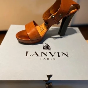 Bruna klackskor från Lanvin - Snygga bruna klackskor från Lanvin med breda remmar och dekorativa knappar. Skorna har en hög klack och är tillverkade i skinn, vilket ger en elegant look.