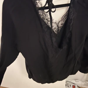 Svart blus med spetsdetaljer - Elegant svart blus med v-ringning och spetsdetaljer. Långärmad design med knytband i nacken. Perfekt för en stilren look.