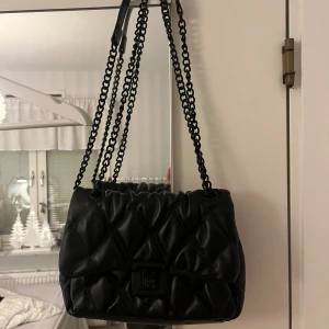 Svart quiltad axelväska - Snygg svart axelväska med quiltad design och kedjeaxelremmar. Perfekt för att ge en stilren touch till din outfit. Väskan har en praktisk storlek och en elegant stängning framtill.