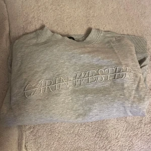 Grå sweatshirt från Carin Wester - Säljer en stilren grå sweatshirt från Carin Wester med broderad logga på bröstet. Tröjan har långa ärmar och en klassisk rund halsringning. Perfekt för en avslappnad look.