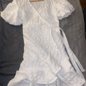 Vit omlottklänning från Pieces - Säljer en söt vit omlottklänning från Pieces med korta puffärmar och knytband i midjan. Klänningen har ett fint blommigt mönster och volangdetaljer längs fållen. Perfekt för sommarens alla tillfällen!
