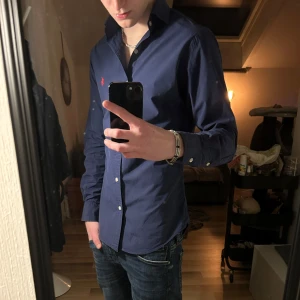Mörkblå Ralph Lauren skjorta - Snygg mörkblå skjorta från Ralph Lauren i slim fit. Skjortan har långa ärmar och knappar framtill. Den är dekorerad med en röd broderad logotyp på bröstet. Perfekt för en stilren look. Skick 8/10