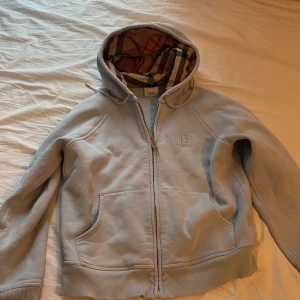 Blå huvtröja från Burberry - Säljer en stilren ljus blå huvtröja från Burberry med dragkedja och klassiskt rutigt foder i huvan. Tröjan har ribbade muddar och två fickor framtill. Perfekt för en avslappnad och trendig look. Kvittot följer ej med. Köpt på en outlet i Paris så den är garanterad äkta. Väldigt konstig storlek så skulle säga att den passar som S även om det står M. Hör av er vid minsta fundering. Pris kan diskuteras!