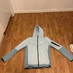 Ljusblå hoodie från Nike - Säljer en ljusblå hoodie från Nike med dragkedja och huva. Jackan har svarta detaljer och en vit Nike-logga på bröstet. Perfekt för en sportig look. Dragkedjan på benet är ihopsydd. Annars är det bra skick. Hela setet för 1499kr, bara hoodie för 800, medans byxor blir 700.