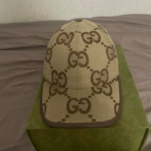 Gucci jumbo keps - Snygg beige keps från Gucci med det ikoniska GG-mönstret i brunt. Kepsen har en klassisk design med rundad skärm och justerbar passform. Perfekt för att ge din outfit en lyxig touch. Självklart är kepsen äkta och allt på bilderna medföljer.