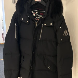 Moose Knuckles Vinterjacka  - Svart herr dunjacka i en smal passform från det kanadensiska märket Moose Knuckles. mjuk, svart naturpäls fastsydd på luvan. Vinterjackan är välvårdad och i mycket bra skick, köptes 2020 hos Johnells så kvitto finns och det är storlek XL på jackan.