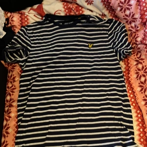 Randig t-shirt från Lyle & Scott - Säljer en snygg randig t-shirt från Lyle & Scott i marinblått och vitt. T-shirten har korta ärmar och ett gult emblem på bröstet. Perfekt för en avslappnad stil.