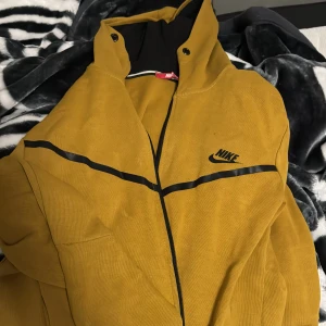 Gul jacka från Nike - Säljer en gul jacka från Nike med svarta detaljer och dragkedja. Jackan har en sportig look med huva och är perfekt för en casual stil. Den har en svart Nike-logga på bröstet.