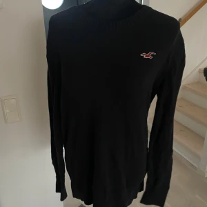 Svart tröja från Hollister - Snygg svart tröja från Hollister med deras ikoniska logga på bröstet. Tröjan har långa ärmar och en ribbad krage som ger en stilren look. Perfekt för en avslappnad stil.