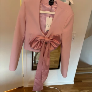 Rosa kavaj med stor rosett från ZARA - Säljer en elegant rosa kavaj från ZARA med en stor dekorativ rosett framtill. Kavajen har en v-ringad design och långa ärmar, perfekt för en stilren look. Materialet är mjukt och ger en lyxig känsla.