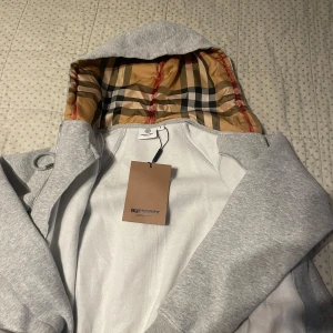 Grå huvtröja från Burberry - Snygg grå huvtröja från Burberry med klassiskt rutigt mönster i huvan. Tröjan har dragkedja framtill och långa ärmar. Perfekt för en stilren och bekväm look.