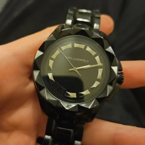 Svart klocka från Karl Lagerfeld - Snygg svart klocka från Karl Lagerfeld med en unik design. Klockan har en stilren urtavla med geometriska detaljer och ett matchande armband i svart. Perfekt för den som vill ha en elegant accessoar.