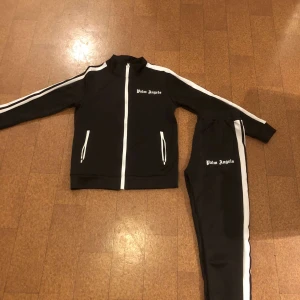 Svart träningsset från Palm Angels - Snyggt svart träningsset från Palm Angels med vita detaljer. Jackan har dragkedja och vita ränder längs ärmarna. Byxorna har matchande vita ränder längs sidorna och elastisk midja för bekväm passform.