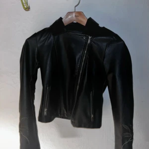 Svart skinnjacka med blommigt broderi - Snygg svart skinnjacka med unikt blommigt broderi på ärmarna. Jackan har en asymmetrisk dragkedja och två fickor med dragkedjor. Perfekt för att ge din outfit en edgy touch.