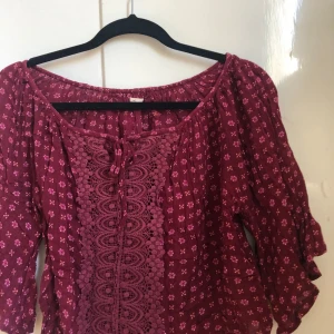 Vinröd Blus från HOLLISTER - Säljer min favorita blus från Hollister med små vita blommor och vackra spetsdetaljer framtill. Blusen har trekvartsärmar och en lös passform, perfekt för vår och sommar. Fint skick och behagligt material 